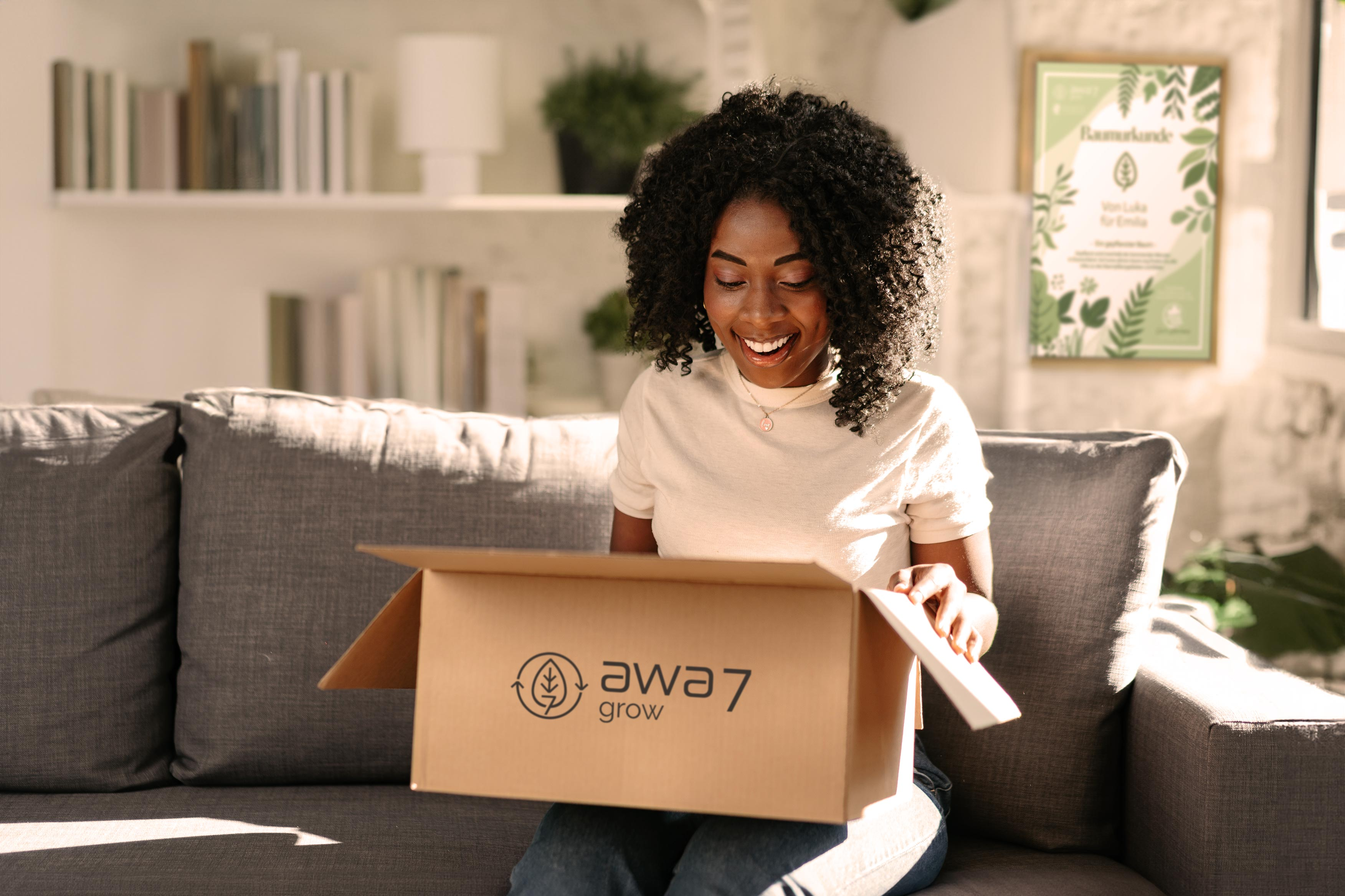 Eine Frau sitzt auf einer grauen Couch und packt freudestrahlend ein awa7 Grow-Paket aus. Im Hintergrund hängt eine awa7-Baumurkunde.
